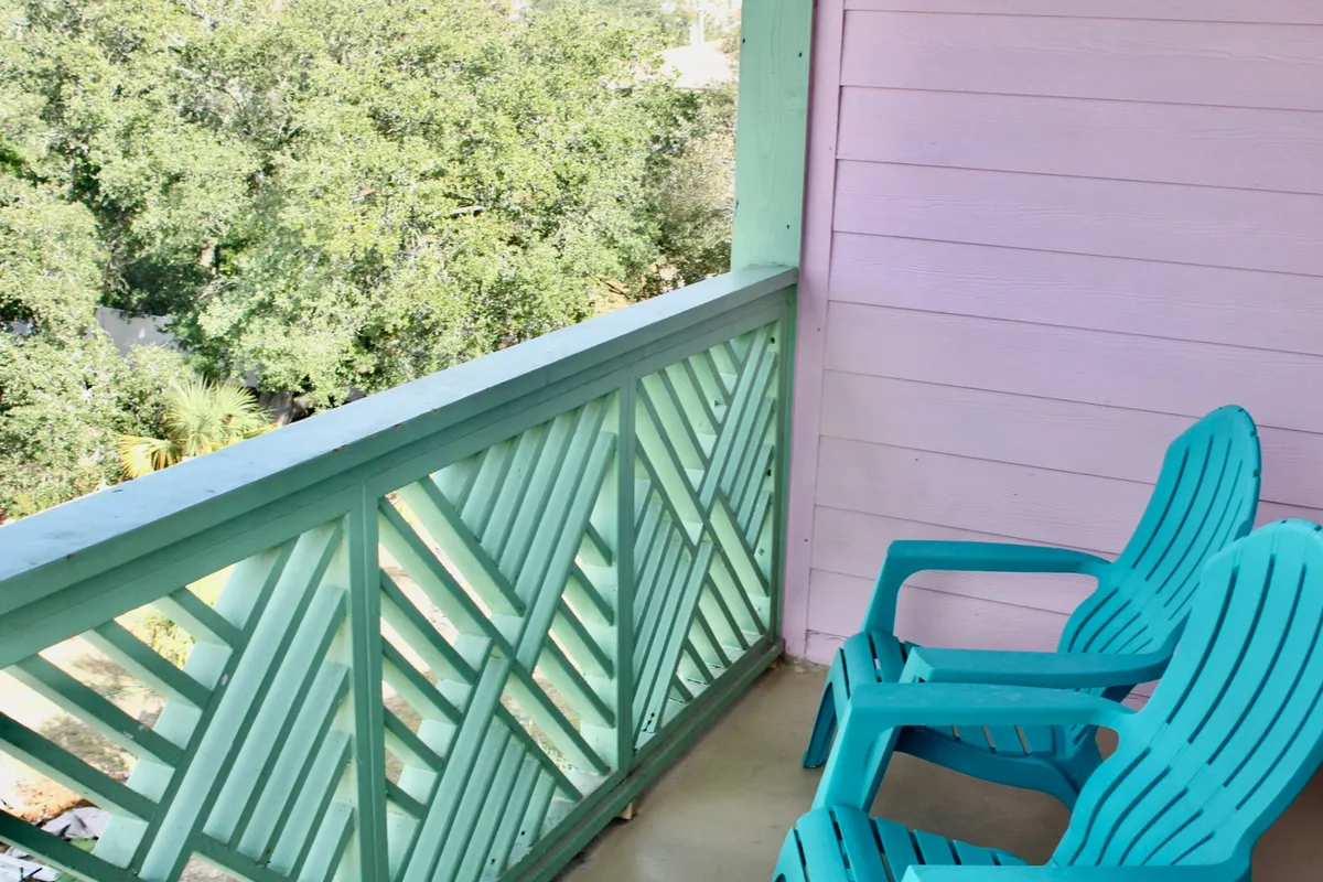 The hero image showcases a tranquil exterior balcony adorned with eye-catching turquoise chairs and a cheerful green railing against a playful pink wall. The inviting setup suggests a perfect spot for sipping coffee, reading a book, or simply enjoying the coastal breeze, with glimpses of the ocean just steps away.